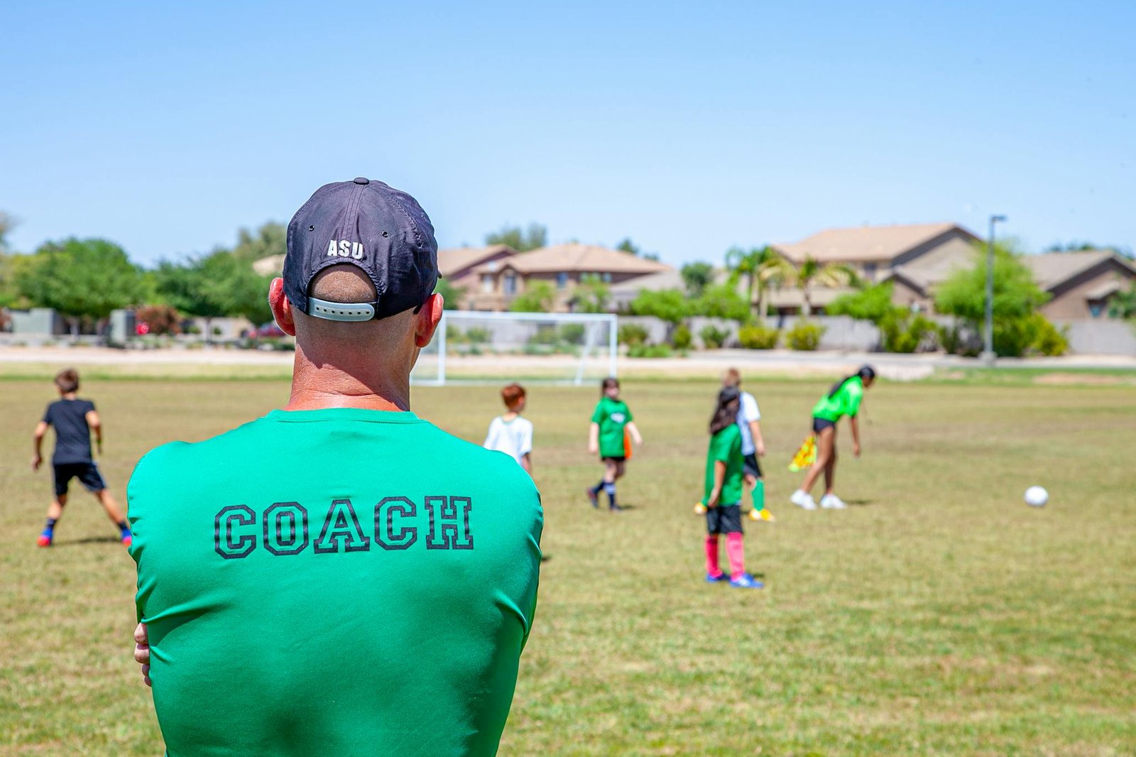 A soccer coach in a cap watches over children practicing on a sunny day at an outdoor field.