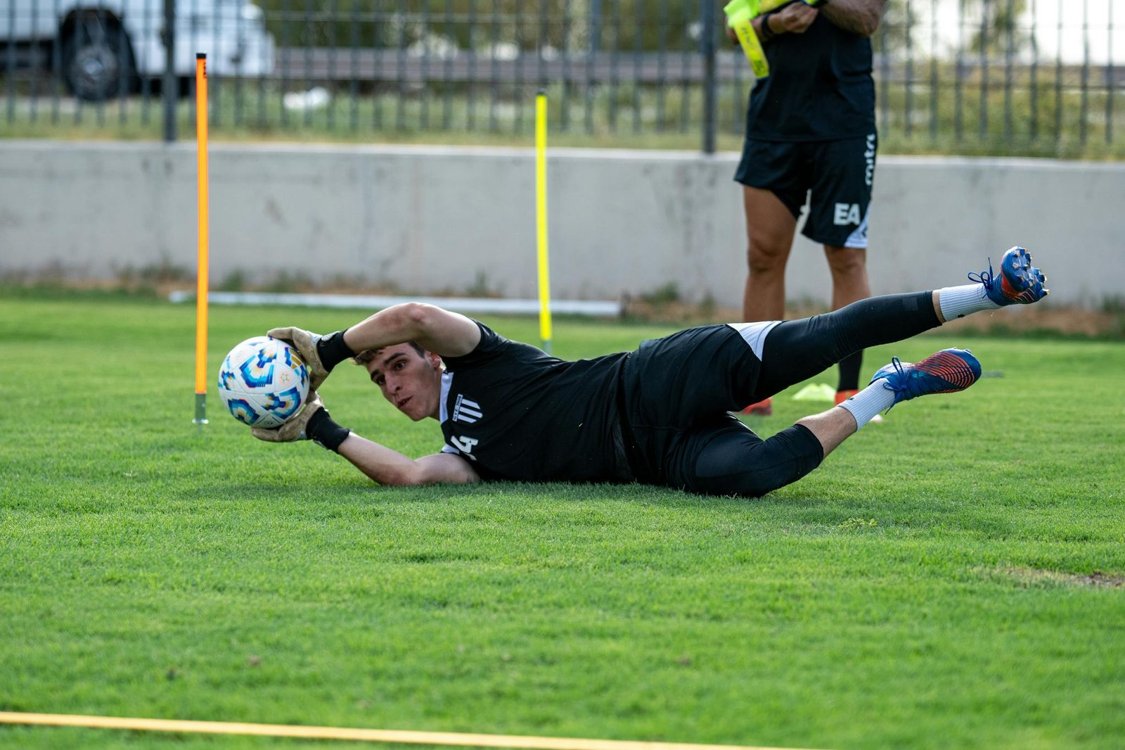 A young soccer goalkeeper dives to catch a ball during training on a grassy field.